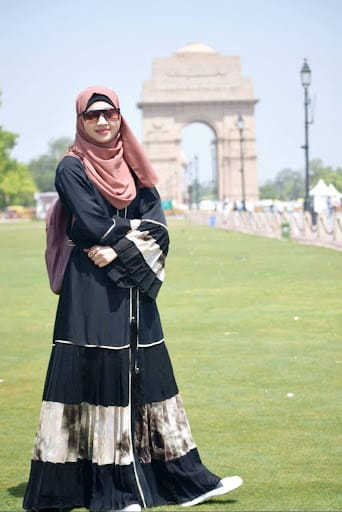 Farhana, wearing a pink hijab, sunglasses, and a long black and white dress, standing in front of a large archway.