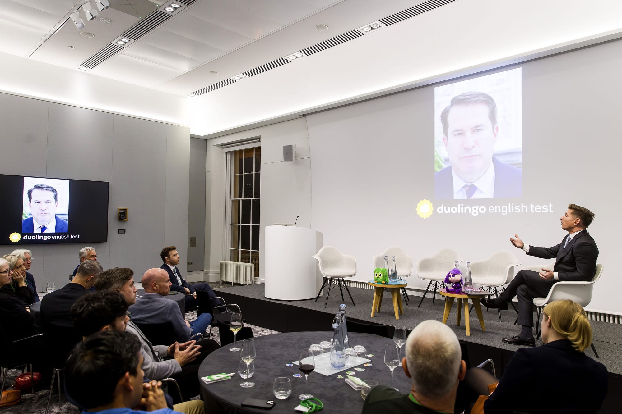 A man on a stage, speaking to another man who is zooming into a panel; his face appears on screen. 