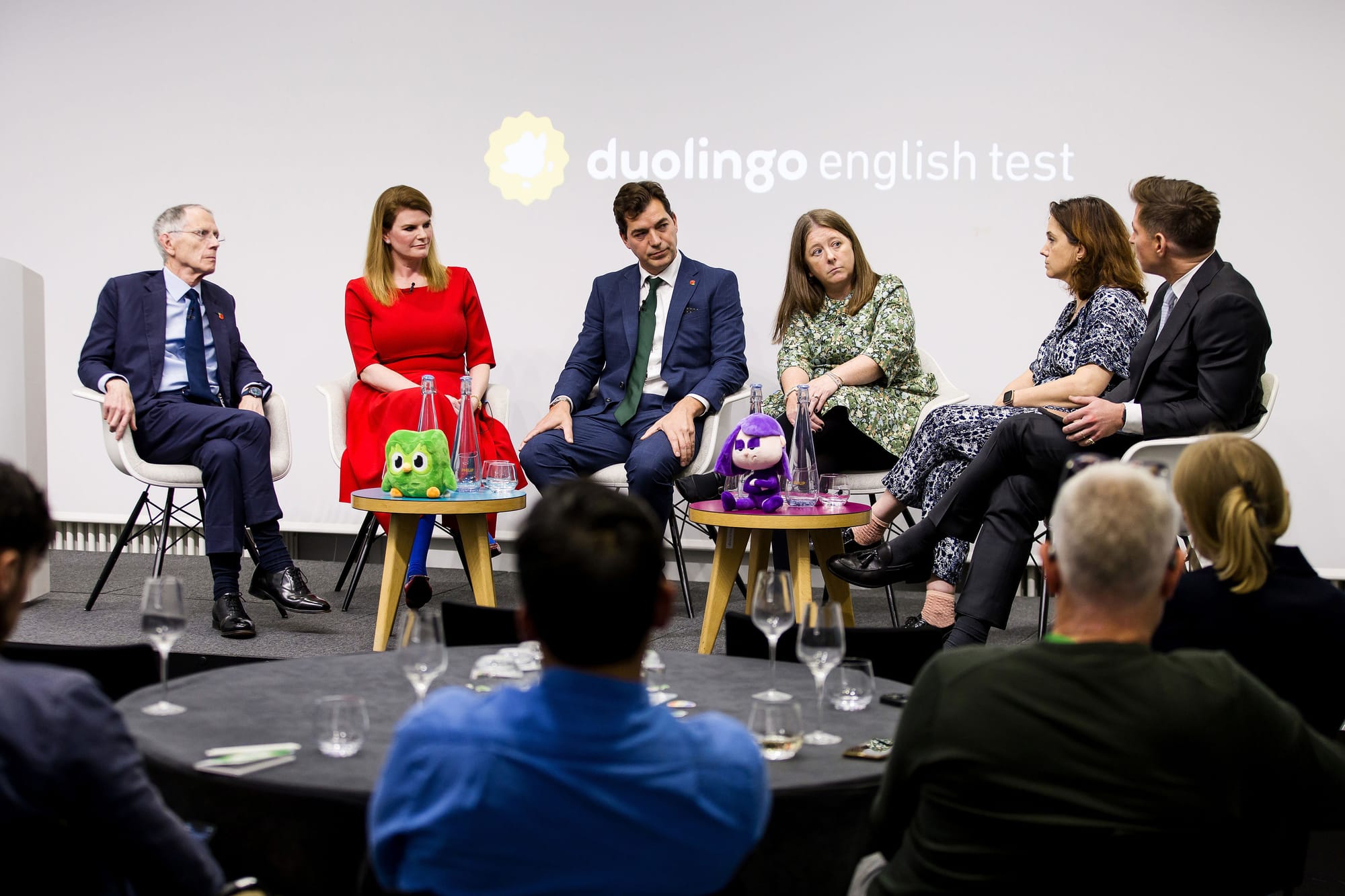 Six people seated on a stage, engaged in conversation; the words "Duolingo English Test" are projected on the wall behind them. 