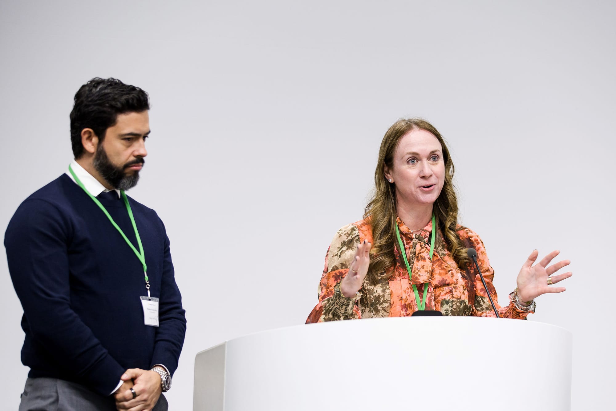 A man stands on the left listening to a woman on the right, speaking from behind a podium