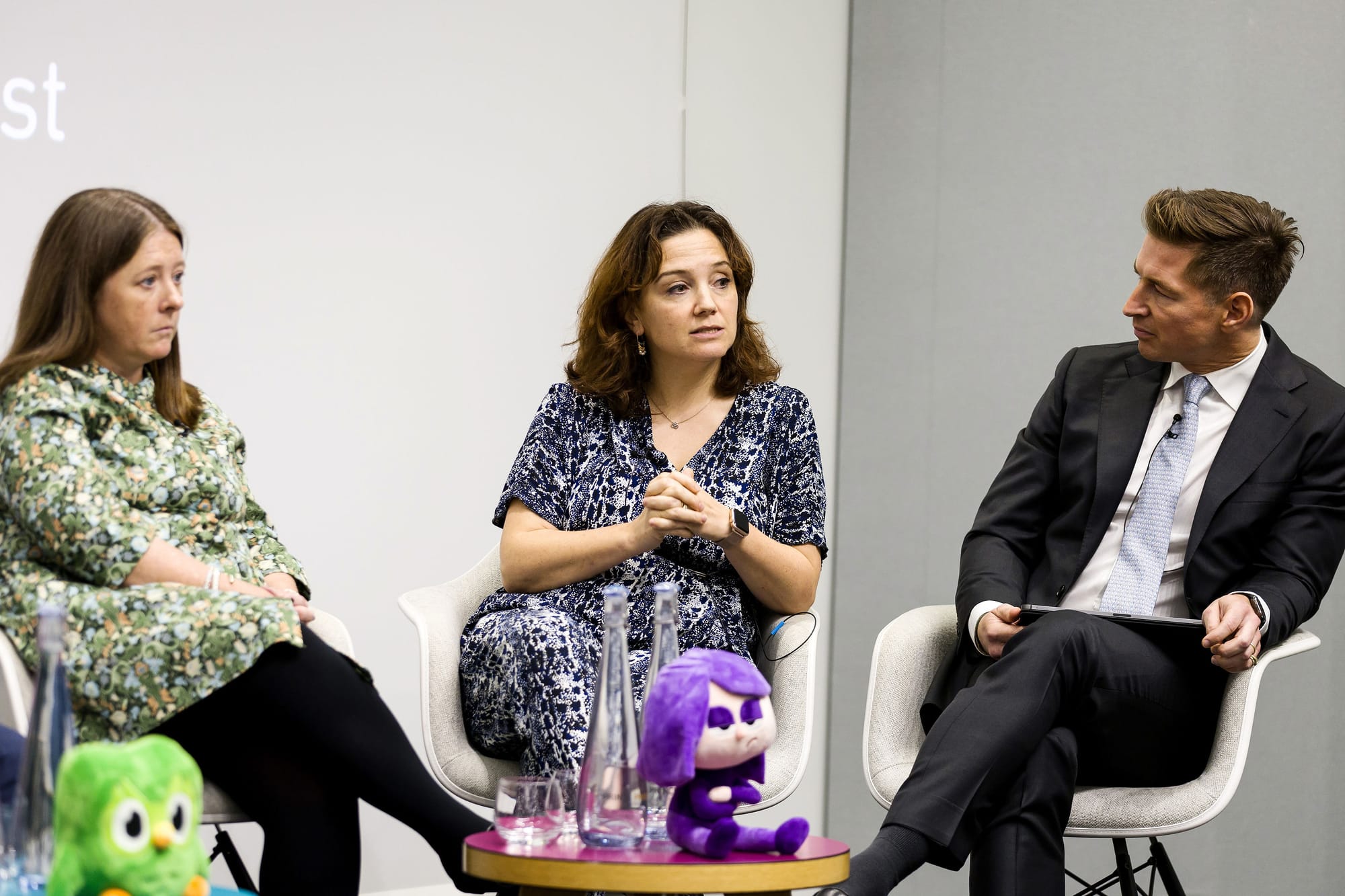 Three people seated on a stage; the woman in the middle is speaking. 