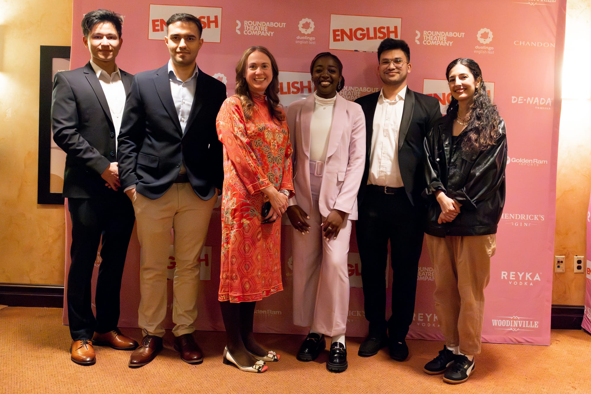 Six people standing in a line, smiling at the camera. Behind them is a pink step-and-repeat printed with the words "ENGLISH" and "Duolingo English Test"