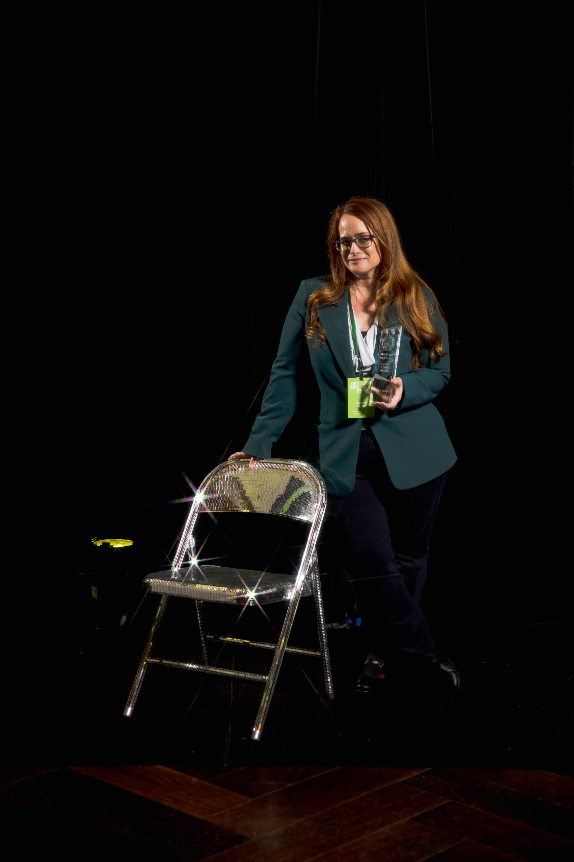 A woman poses against a black backdrop. She is leaning with one hand against a folding chair and holding a We Rise Together award in the other hand.